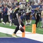 FootballR - NFL Alvin Kamara - Ein Spieler der New Orleans Saints, der eine schwarz-goldene Uniform mit der Nummer 41 trägt, überquert mit einem Football die Endzone. Die Endzone hat einen grünen Rasen und eine blaue Umrandung. Im Hintergrund sind Zuschauer, Teams und ein Kamerateam zu sehen, die das Spielgeschehen einfangen. Diese Beschreibung wurde automatisch generiert.