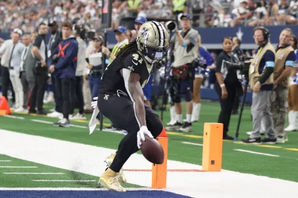 FootballR - NFL Alvin Kamara - Ein Spieler der New Orleans Saints, der eine schwarz-goldene Uniform mit der Nummer 41 trägt, überquert mit einem Football die Endzone. Die Endzone hat einen grünen Rasen und eine blaue Umrandung. Im Hintergrund sind Zuschauer, Teams und ein Kamerateam zu sehen, die das Spielgeschehen einfangen. Diese Beschreibung wurde automatisch generiert.