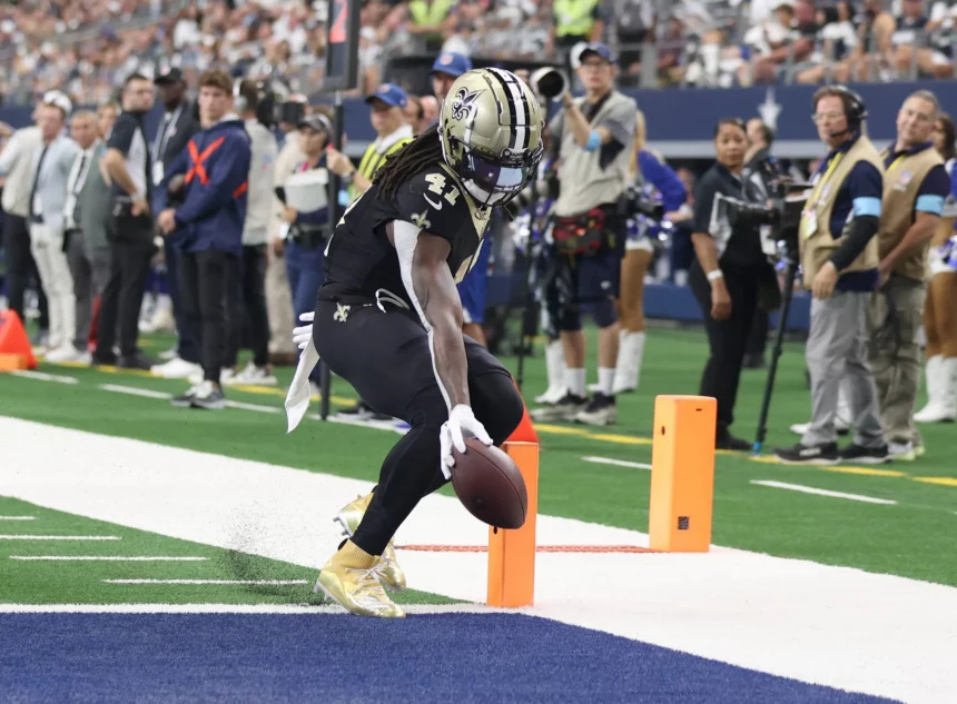 FootballR - NFL Alvin Kamara - Ein Spieler der New Orleans Saints, der eine schwarz-goldene Uniform mit der Nummer 41 trägt, überquert mit einem Football die Endzone. Die Endzone hat einen grünen Rasen und eine blaue Umrandung. Im Hintergrund sind Zuschauer, Teams und ein Kamerateam zu sehen, die das Spielgeschehen einfangen. Diese Beschreibung wurde automatisch generiert.