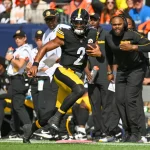 FootballR - NFL - Ein Footballspieler in einer schwarz-gelben Uniform mit der Nummer 2, Justin Fields, läuft während eines Spiels mit dem Ball. Trainer und andere Spieler sind an der Seitenlinie zu sehen, einige mit unterstützenden Gesten. Im Hintergrund beobachten Zuschauer von den Tribünen aus das Spiel. Auch die Spieler der gegnerischen Mannschaft sind zu sehen. Diese Beschreibung wurde automatisch generiert.