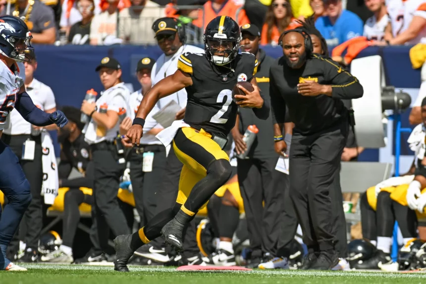 FootballR - NFL - Ein Footballspieler in einer schwarz-gelben Uniform mit der Nummer 2, Justin Fields, läuft während eines Spiels mit dem Ball. Trainer und andere Spieler sind an der Seitenlinie zu sehen, einige mit unterstützenden Gesten. Im Hintergrund beobachten Zuschauer von den Tribünen aus das Spiel. Auch die Spieler der gegnerischen Mannschaft sind zu sehen. Diese Beschreibung wurde automatisch generiert.