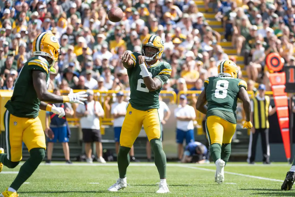 FootballR - NFL - Ein Footballspieler in einem grün-gelben Trikot der Green Bay Packers, Nummer 2, wirft während des Spiels den Ball zu einem Mitspieler mit der Nummer 86. Ein weiterer Mitspieler, Nummer 8, läuft in der Nähe. Das Stadion ist voller Zuschauer, viele in den grün-gelben Trikots von Malik Willis und Jordan Love, die gespannt zuschauen. Diese Beschreibung wurde automatisch generiert.