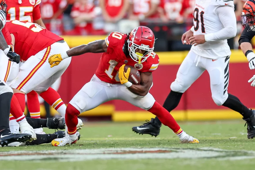 FootballR - NFL - Isiah Pacheco, der Running Back der Chiefs, trägt eine rote Uniform, weiße Hosen und gelbe Handschuhe, während er mit dem Ball nach vorne stürmt. Er wird von Spielern verfolgt, die weiße Uniformen mit schwarzen und orangefarbenen Details tragen. Die spannende Action entfaltet sich auf einem belebten Footballfeld mit einer aufgeregten Menge im Hintergrund. Diese Beschreibung wurde automatisch generiert.