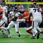 FootballR - NFL - Ein Footballspieler in weißer Uniform läuft mit dem Ball, was an Joe Mixon erinnert, während er von mehreren Gegenspielern in orangefarbenen Uniformen verfolgt wird. Ein Spieler in orangefarbener Uniform streckt die Hand aus, um den Ballträger zu tackeln. Das Spiel findet auf einem gut beleuchteten Feld mit einer verschwommenen Menschenmenge im Hintergrund statt. Diese Beschreibung wurde automatisch generiert.