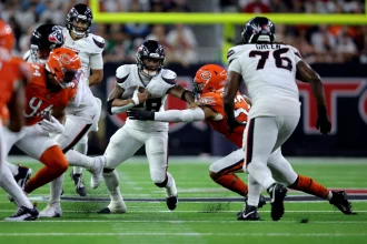 FootballR - NFL - Ein Footballspieler in weißer Uniform läuft mit dem Ball, was an Joe Mixon erinnert, während er von mehreren Gegenspielern in orangefarbenen Uniformen verfolgt wird. Ein Spieler in orangefarbener Uniform streckt die Hand aus, um den Ballträger zu tackeln. Das Spiel findet auf einem gut beleuchteten Feld mit einer verschwommenen Menschenmenge im Hintergrund statt. Diese Beschreibung wurde automatisch generiert.