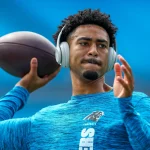 FootballR - NFL - Ein Footballspieler in einem blauen Shirt mit dem Logo der Carolina Panthers wärmt sich auf dem Feld auf. Bryce Young trägt große silberne Kopfhörer, hält einen Football in seiner rechten Hand und scheint sich mitten in einer Wurfbewegung zu befinden. Im Hintergrund sind blaue Stadionsitze zu sehen. Diese Beschreibung wurde automatisch generiert.