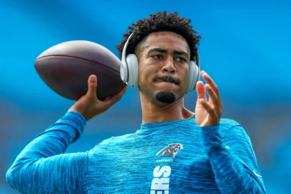 FootballR - NFL - Ein Footballspieler in einem blauen Shirt mit dem Logo der Carolina Panthers wärmt sich auf dem Feld auf. Bryce Young trägt große silberne Kopfhörer, hält einen Football in seiner rechten Hand und scheint sich mitten in einer Wurfbewegung zu befinden. Im Hintergrund sind blaue Stadionsitze zu sehen. Diese Beschreibung wurde automatisch generiert.