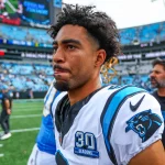 FootballR - NFL - Ein Footballspieler mit kurzen Locken und einem weißen Carolina Panthers-Trikot steht konzentriert auf dem Feld. Bryce Young zieht die Aufmerksamkeit auf sich, während das Stadion im Hintergrund voller Zuschauer den Atem anzuhalten scheint. Andere Personen sind zu sehen, darunter ein Mann in einem schwarzen Trikot. Diese Beschreibung wurde automatisch generiert.