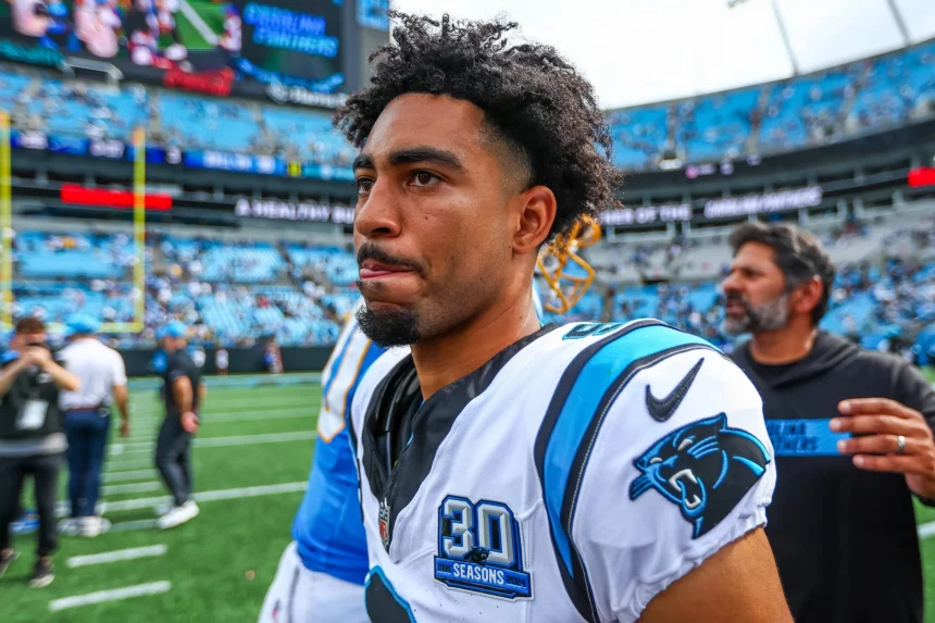 FootballR - NFL - Ein Footballspieler mit kurzen Locken und einem weißen Carolina Panthers-Trikot steht konzentriert auf dem Feld. Bryce Young zieht die Aufmerksamkeit auf sich, während das Stadion im Hintergrund voller Zuschauer den Atem anzuhalten scheint. Andere Personen sind zu sehen, darunter ein Mann in einem schwarzen Trikot. Diese Beschreibung wurde automatisch generiert.