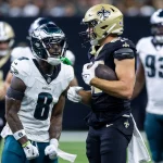 FootballR - NFL - Ein Spieler der New Orleans Saints, in schwarzer Uniform und mit goldenem Helm, steht mit einem Football in der Hand da. Ein wütender Spieler der Philadelphia Eagles, Nummer 8, steht ihm gegenüber und schreit. Unter den Spielern auf dem Feld ist C.J. Gardner-Johnson, der ebenfalls von der Intensität des Augenblicks erfasst ist. Diese Beschreibung wurde automatisch generiert.