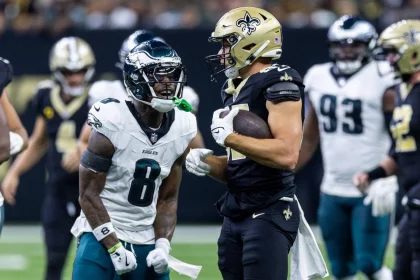 FootballR - NFL - Ein Spieler der New Orleans Saints, in schwarzer Uniform und mit goldenem Helm, steht mit einem Football in der Hand da. Ein wütender Spieler der Philadelphia Eagles, Nummer 8, steht ihm gegenüber und schreit. Unter den Spielern auf dem Feld ist C.J. Gardner-Johnson, der ebenfalls von der Intensität des Augenblicks erfasst ist. Diese Beschreibung wurde automatisch generiert.