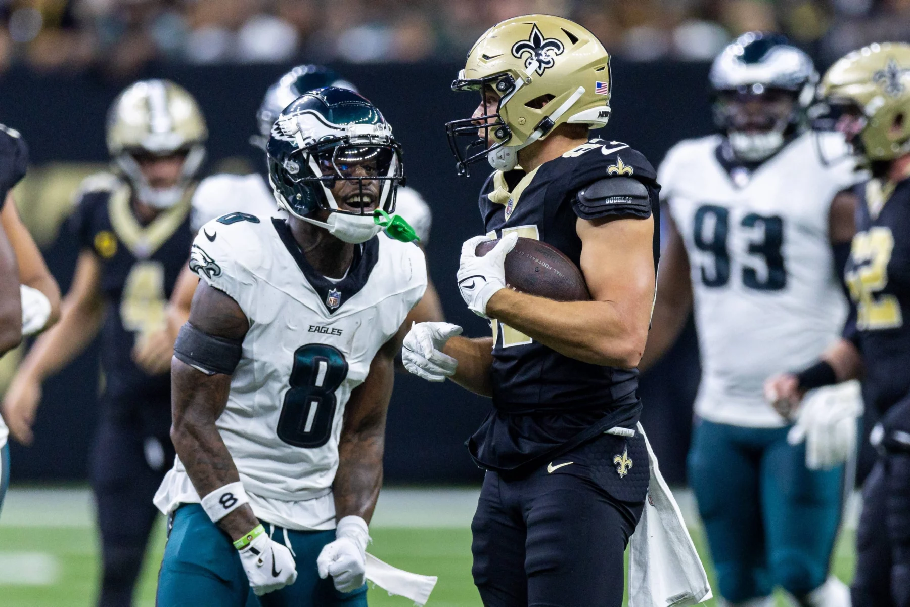 FootballR - NFL - Ein Spieler der New Orleans Saints, in schwarzer Uniform und mit goldenem Helm, steht mit einem Football in der Hand da. Ein wütender Spieler der Philadelphia Eagles, Nummer 8, steht ihm gegenüber und schreit. Unter den Spielern auf dem Feld ist C.J. Gardner-Johnson, der ebenfalls von der Intensität des Augenblicks erfasst ist. Diese Beschreibung wurde automatisch generiert.