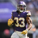 FootballR - NFL - Ein Footballspieler, Aaron Jones, trägt eine lila-weiße Uniform mit der Nummer 33 auf dem Trikot und läuft während eines Spiels über das Feld. Er hält einen Football in seinem rechten Arm und trägt einen Helm mit Gesichtsmaske. Der Spieler trägt gelbe Handschuhe und im Hintergrund ist eine verschwommene Menschenmenge zu sehen. Diese Beschreibung wurde automatisch generiert.