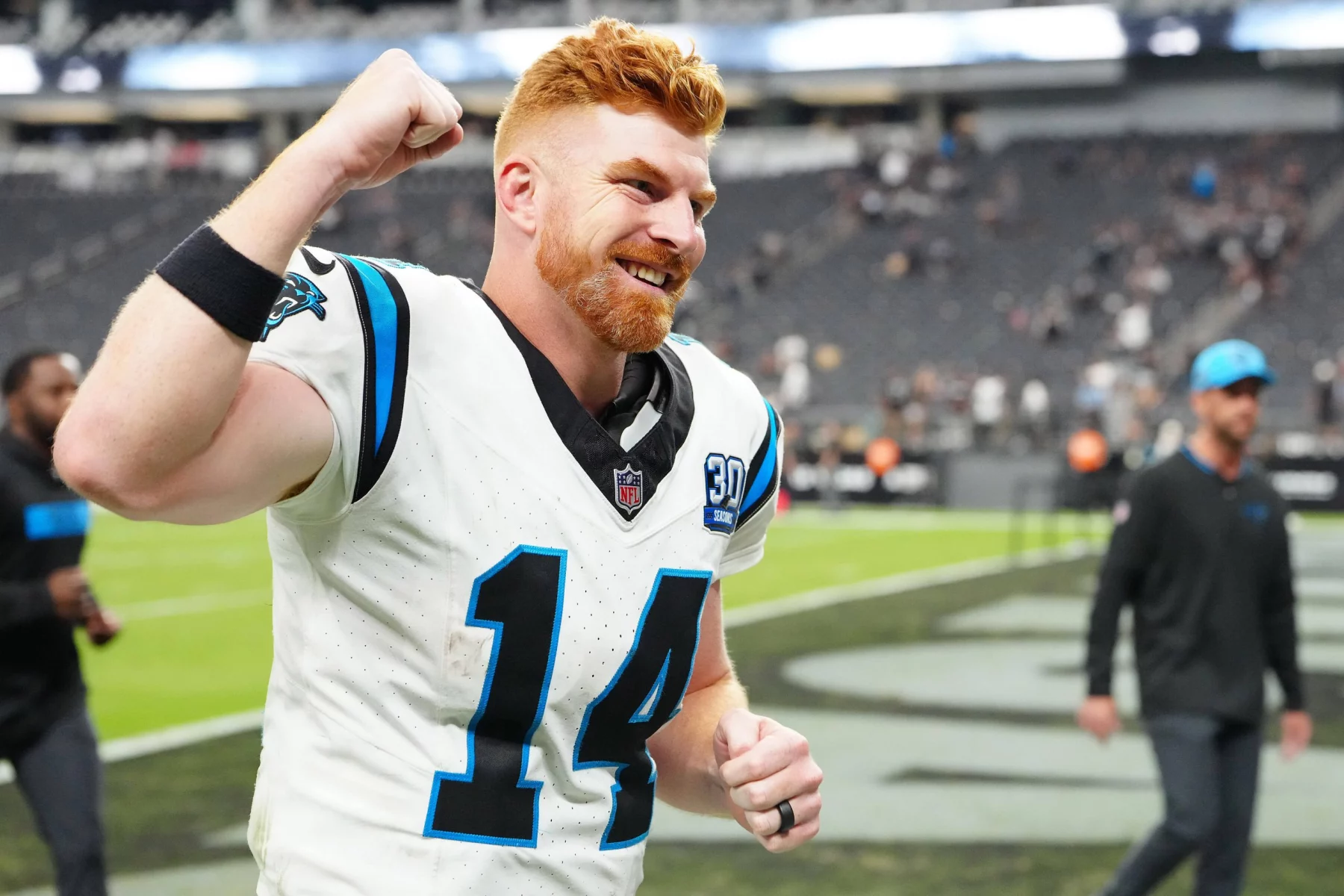 Der Quarterback Wechsel der Carolina Panthers machte sich bezahlt. Andy Dalton trägt ein weißes Carolina Panthers-Trikot mit der Nummer 14 und streckt jubelnd seine rechte Faust in die Höhe. Das Trikot hat einen schwarz-blauen Rand und der Spieler scheint auf einem Feld in einem Stadion zu stehen, im Hintergrund sind andere Menschen zu sehen.