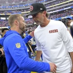 Aufeinandertreffen in Australien - Die Rams bezwangen überraschend die 49ers in letzter Sekunde. Zwei Footballtrainer geben sich nach dem Spiel auf dem Spielfeld die Hand. Der Trainer links - Sean McVay - trägt einen blauen Kapuzenpullover mit dem Logo der Los Angeles Rams auf dem Ärmel. Der Trainer rechts - Kyle Shanahan - trägt ein weißes Hemd mit dem Aufdruck „49ers“ und eine schwarze Kappe. Im Hintergrund des Stadions dieses NFL-Events sind Kamerateam und anderes Personal zu sehen.