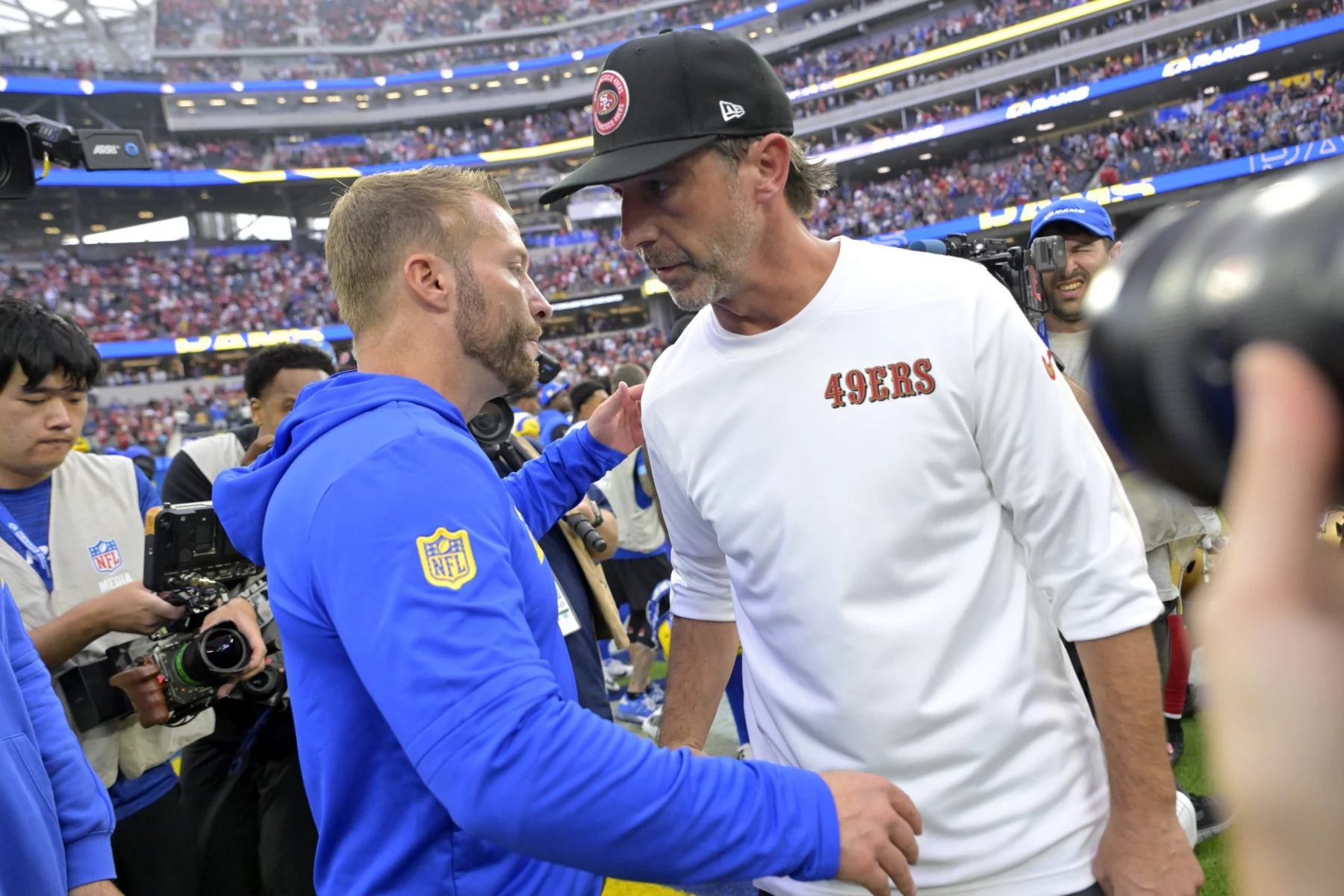 Aufeinandertreffen in Australien - Die Rams bezwangen überraschend die 49ers in letzter Sekunde. Zwei Footballtrainer geben sich nach dem Spiel auf dem Spielfeld die Hand. Der Trainer links - Sean McVay - trägt einen blauen Kapuzenpullover mit dem Logo der Los Angeles Rams auf dem Ärmel. Der Trainer rechts - Kyle Shanahan - trägt ein weißes Hemd mit dem Aufdruck „49ers“ und eine schwarze Kappe. Im Hintergrund des Stadions dieses NFL-Events sind Kamerateam und anderes Personal zu sehen.