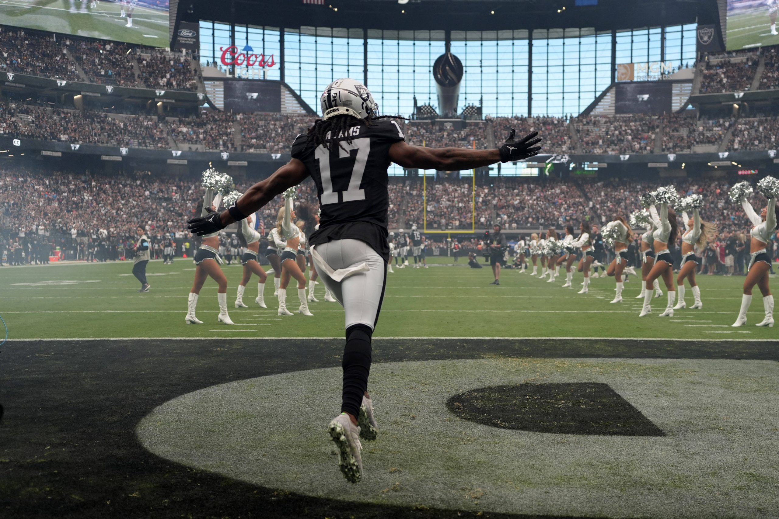 FootballR - NFL - Ein Footballspieler in schwarz-weißer Uniform mit der Nummer 17 läuft mit ausgestreckten Armen über das Feld. Im Hintergrund stellen sich Cheerleader in der Nähe des Publikums in einem voll besetzten Stadion in Las Vegas auf. Auf der großen Leinwand wird der Spielverlauf übertragen, und unter dem Verletzten Davante Adams ist eine Werbung von Coors zu sehen. Diese Beschreibung wurde automatisch generiert.