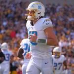 FootballR - NFL - Ein Footballspieler in weiß-blauer Uniform mit der Nummer 10 auf seinem Trikot steht auf dem Feld. Er trägt einen Helm und ein Playbook-Armband und sieht aus wie Justin Herbert. Seine Teamkollegen, ebenfalls in weiß-blauen Uniformen, sind in der Nähe. Das Stadion ist voller Zuschauer und im Hintergrund ist ein Schiedsrichter zu sehen. Diese Beschreibung wurde automatisch generiert.