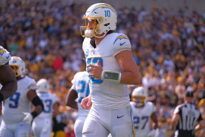 FootballR - NFL - Ein Footballspieler in weiß-blauer Uniform mit der Nummer 10 auf seinem Trikot steht auf dem Feld. Er trägt einen Helm und ein Playbook-Armband und sieht aus wie Justin Herbert. Seine Teamkollegen, ebenfalls in weiß-blauen Uniformen, sind in der Nähe. Das Stadion ist voller Zuschauer und im Hintergrund ist ein Schiedsrichter zu sehen. Diese Beschreibung wurde automatisch generiert.