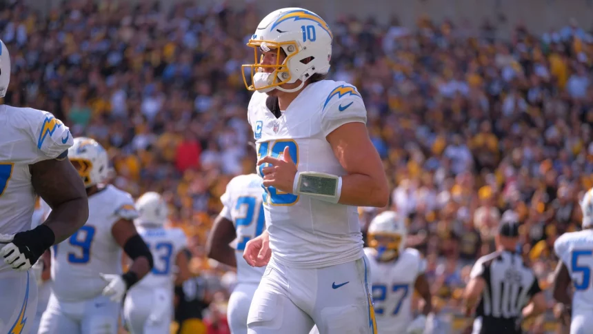 FootballR - NFL - Ein Footballspieler in weiß-blauer Uniform mit der Nummer 10 auf seinem Trikot steht auf dem Feld. Er trägt einen Helm und ein Playbook-Armband und sieht aus wie Justin Herbert. Seine Teamkollegen, ebenfalls in weiß-blauen Uniformen, sind in der Nähe. Das Stadion ist voller Zuschauer und im Hintergrund ist ein Schiedsrichter zu sehen. Diese Beschreibung wurde automatisch generiert.