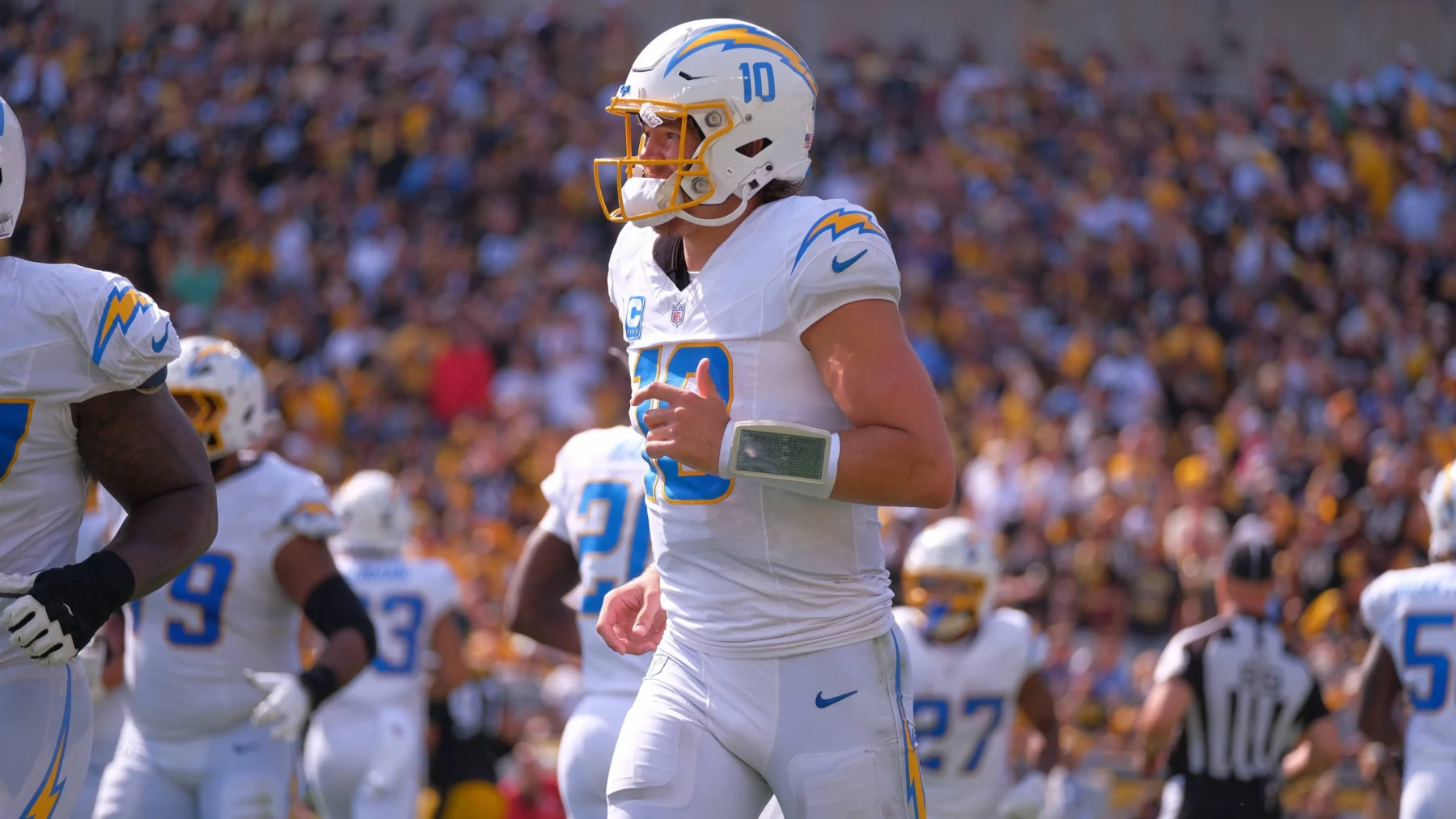 FootballR - NFL - Ein Footballspieler in weiß-blauer Uniform mit der Nummer 10 auf seinem Trikot steht auf dem Feld. Er trägt einen Helm und ein Playbook-Armband und sieht aus wie Justin Herbert. Seine Teamkollegen, ebenfalls in weiß-blauen Uniformen, sind in der Nähe. Das Stadion ist voller Zuschauer und im Hintergrund ist ein Schiedsrichter zu sehen. Diese Beschreibung wurde automatisch generiert.