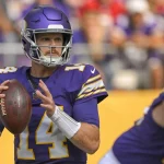 FootballR - NFL - Ein Footballspieler in lila-weißer Uniform mit der Nummer 14 auf seinem Trikot, der an Sam Darnold erinnert, hält den Football und bereitet sich auf einen Pass vor. Er trägt einen lila Helm mit Gesichtsschutz und im Hintergrund sind andere Spieler und verschwommene Zuschauer zu sehen. Diese Beschreibung wurde automatisch generiert.