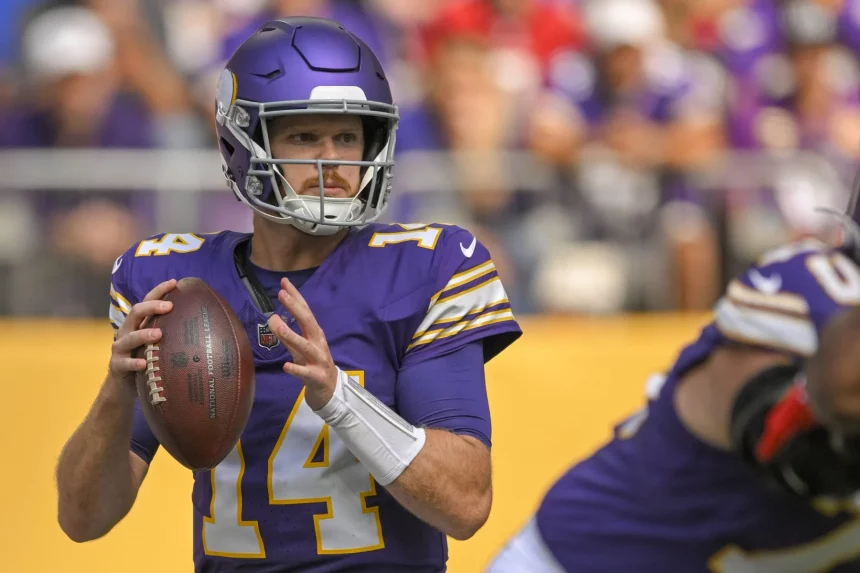 FootballR - NFL - Ein Footballspieler in lila-weißer Uniform mit der Nummer 14 auf seinem Trikot, der an Sam Darnold erinnert, hält den Football und bereitet sich auf einen Pass vor. Er trägt einen lila Helm mit Gesichtsschutz und im Hintergrund sind andere Spieler und verschwommene Zuschauer zu sehen. Diese Beschreibung wurde automatisch generiert.