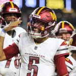 FootballR - DAZN NFL Programm Woche 6 - Spielverschiebungen - NFL Regular Season 2024 - Ein Footballspieler im weißen „Washington Commanders“-Trikot mit der Nummer 5, vermutlich Jayden Daniels, lächelt und hebt die Hand. Er ist von Teamkollegen in ähnlichen Uniformen umgeben, auf deren Helmen das Logo des Teams zu sehen ist. Im Hintergrund ist ein Teil einer Stadionkulisse zu sehen. Diese Beschreibung wurde automatisch generiert.