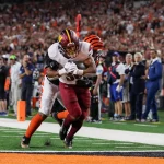 FootballR - NFL - Austin Ekeler erzielt einen Touchdown. Zwei American-Football-Spieler in Uniform sind mitten im Spiel auf einem Feld. Ein Spieler, in einer weißen Commanders-Uniform mit kastanienbraunen und gelben Akzenten, hält den Ball fest, während der andere Spieler, in einer schwarz-orange gestreiften Cardinals-Uniform, ihn von hinten angreift. Eine Menge Zuschauer und Offizielle stehen an der Seitenlinie. Diese Beschreibung wurde automatisch generiert.