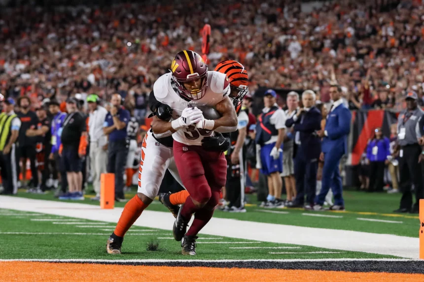 FootballR - NFL - Austin Ekeler erzielt einen Touchdown. Zwei American-Football-Spieler in Uniform sind mitten im Spiel auf einem Feld. Ein Spieler, in einer weißen Commanders-Uniform mit kastanienbraunen und gelben Akzenten, hält den Ball fest, während der andere Spieler, in einer schwarz-orange gestreiften Cardinals-Uniform, ihn von hinten angreift. Eine Menge Zuschauer und Offizielle stehen an der Seitenlinie. Diese Beschreibung wurde automatisch generiert.
