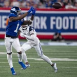 FootballR - NFL - Malik Nabers - Ein Footballspieler in blauer Uniform streckt beide Hände aus, um einen Football in der Luft zu fangen, während Malik Nabers in weißer Uniform versucht, ihn zu blockieren. Sie stehen auf einem Footballfeld, im Hintergrund ist auf einem Banner „New York Football“ zu sehen. Die Zuschauer auf den Tribünen sind unscharf. Diese Beschreibung wurde automatisch generiert.