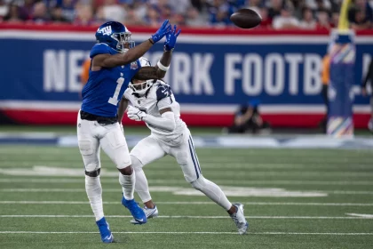 FootballR - NFL - Malik Nabers - Ein Footballspieler in blauer Uniform streckt beide Hände aus, um einen Football in der Luft zu fangen, während Malik Nabers in weißer Uniform versucht, ihn zu blockieren. Sie stehen auf einem Footballfeld, im Hintergrund ist auf einem Banner „New York Football“ zu sehen. Die Zuschauer auf den Tribünen sind unscharf. Diese Beschreibung wurde automatisch generiert.