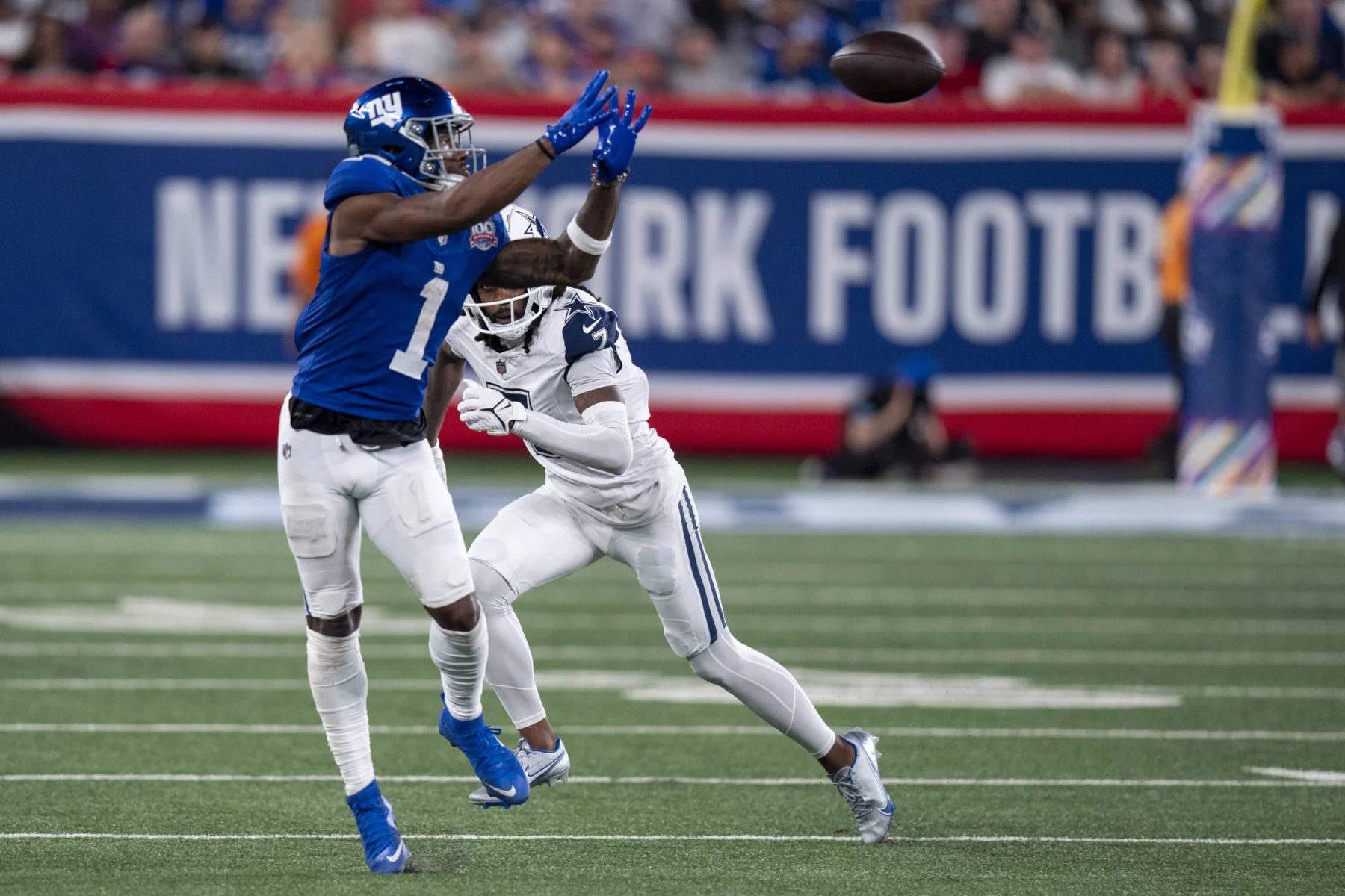 FootballR - NFL - Malik Nabers - Ein Footballspieler in blauer Uniform streckt beide Hände aus, um einen Football in der Luft zu fangen, während Malik Nabers in weißer Uniform versucht, ihn zu blockieren. Sie stehen auf einem Footballfeld, im Hintergrund ist auf einem Banner „New York Football“ zu sehen. Die Zuschauer auf den Tribünen sind unscharf. Diese Beschreibung wurde automatisch generiert.