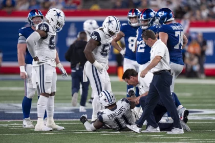 FootballR - NFL - Eine Gruppe von Cowboys- und Giants-Spielern steht um einen verletzten Cowboys-Spieler, Micah Parsons, der mit der Trikotnummer 11 und der Aufschrift „PARSONS“ auf dem Feld liegt. Medizinisches Personal kümmert sich um ihn. Die Szene ist angespannt, die Spieler zeigen sich besorgt. DeMarcus Lawrence beobachtet sie aufmerksam. Es scheint, als ob es sich in einem Stadion abspielt. Diese Beschreibung wurde automatisch generiert.