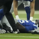 FootballR - NFL - Quarterback Anthony Richardson von den Indianapolis Colts, der ein blaues Trikot mit der Nummer 30 trägt, liegt nach einer Hüftverletzung auf dem Feld. Teamkollegen in weißen und blauen Trikots kümmern sich um ihn, während der Ball in der Nähe liegt. Die Beine eines Schiedsrichters und eines weiteren Spielers in einer gelb-schwarzen Uniform sind ebenfalls zu sehen. Diese Beschreibung wurde automatisch generiert.