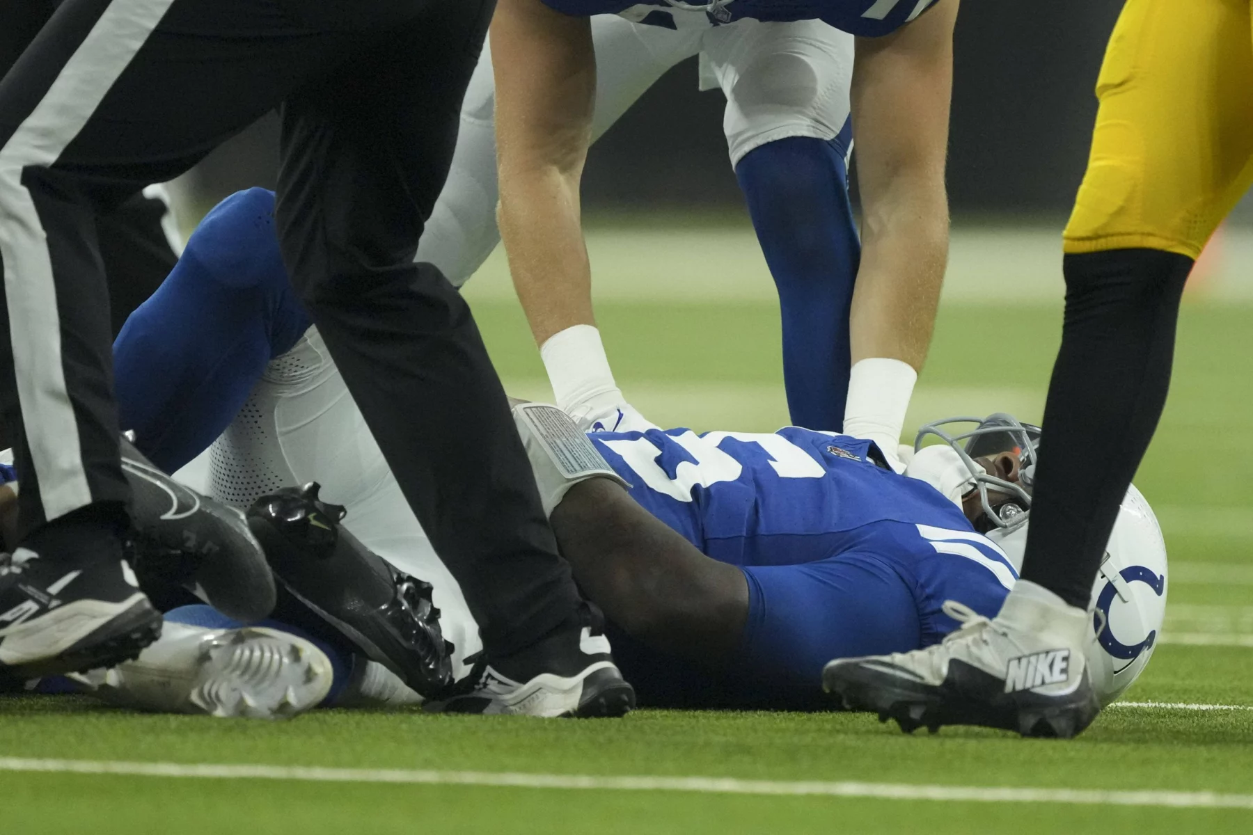 FootballR - NFL - Quarterback Anthony Richardson von den Indianapolis Colts, der ein blaues Trikot mit der Nummer 30 trägt, liegt nach einer Hüftverletzung auf dem Feld. Teamkollegen in weißen und blauen Trikots kümmern sich um ihn, während der Ball in der Nähe liegt. Die Beine eines Schiedsrichters und eines weiteren Spielers in einer gelb-schwarzen Uniform sind ebenfalls zu sehen. Diese Beschreibung wurde automatisch generiert.