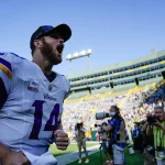 FootballR - NFL - Ein Footballspieler, Sam Darnold, in einem weißen Minnesota Vikings Trikot mit der Nummer 14 schreit aufgeregt am Spielfeldrand eines mit Zuschauern gefüllten Stadions. Unter dem klaren blauen Himmel trägt der Spieler eine schwarze Kappe, während ein Kamerateam den Moment festhält und den 4:0-Saisonstart des Teams feiert. Diese Beschreibung wurde automatisch generiert.