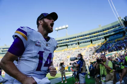 FootballR - NFL - Ein Footballspieler, Sam Darnold, in einem weißen Minnesota Vikings Trikot mit der Nummer 14 schreit aufgeregt am Spielfeldrand eines mit Zuschauern gefüllten Stadions. Unter dem klaren blauen Himmel trägt der Spieler eine schwarze Kappe, während ein Kamerateam den Moment festhält und den 4:0-Saisonstart des Teams feiert. Diese Beschreibung wurde automatisch generiert.