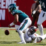 FootballR - NFL - Ein Footballspieler der Philadelphia Eagles in grünen Trikots wird von einem Spieler in weißem Trikot zu Boden gerissen. Jalen Hurts in Grün ist gebeugt und versucht, einen fallengelassenen Football zurückzuholen, während der Spieler in Weiß auf dem Feld liegt und sein Bein hält. Spieler und ein roter Werbehintergrund sind sichtbar. Diese Beschreibung wurde automatisch generiert.