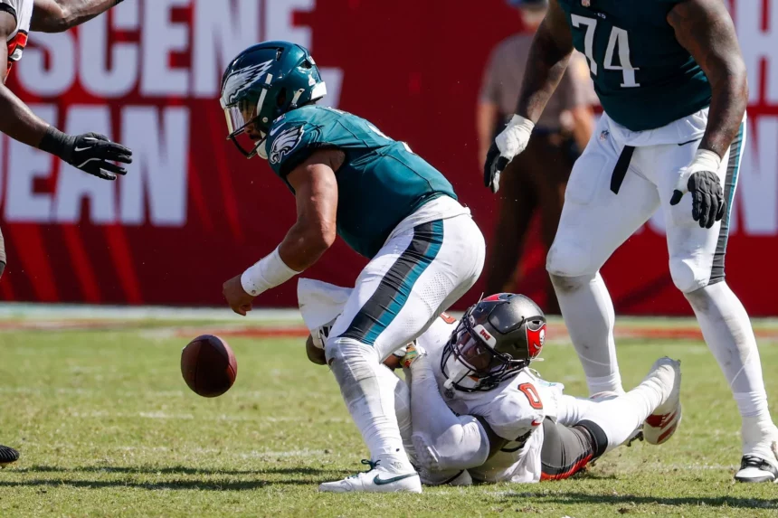 FootballR - NFL - Ein Footballspieler der Philadelphia Eagles in grünen Trikots wird von einem Spieler in weißem Trikot zu Boden gerissen. Jalen Hurts in Grün ist gebeugt und versucht, einen fallengelassenen Football zurückzuholen, während der Spieler in Weiß auf dem Feld liegt und sein Bein hält. Spieler und ein roter Werbehintergrund sind sichtbar. Diese Beschreibung wurde automatisch generiert.
