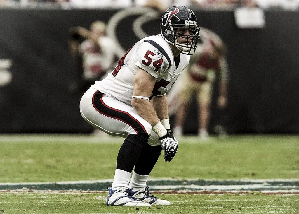 FootballR - NFL - Ein Footballspieler in weißer Houston Texans-Uniform mit der Nummer 54 kauert in Abwehrhaltung auf dem Feld. Er trägt einen Helm mit Gesichtsschutz, Handschuhe, weiße Socken und Stollenschuhe. Im Hintergrund sind verschwommene Figuren und Stadionsitze zu sehen – ein Hauptaugenmerk für alle, die in dieser kommenden Saison NFL-Wetten abschließen. Diese Beschreibung wurde automatisch generiert.