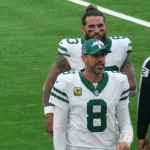 FootballR - NFL - Ein Footballspieler in weiß-grüner New York-Jets-Uniform, Nummer 8, spricht mit einem Schiedsrichter. Er trägt eine Kappe, während ein anderer Jets-Spieler mit langen Haaren und Bart hinter ihm steht. Das Spielfeld ist grün, der Schiedsrichter trägt schwarz-weiße Streifen – fast wie ein automatisch gespeicherter Entwurf. Diese Beschreibung wurde automatisch generiert.