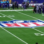 FootballR - NFL Spiel - Footballspieler in lila Trikots versammeln sich auf dem Feld nahe der 50-Yard-Linie. In der Mitte des Spielfelds ist ein auffälliges NFL-Logo aufgemalt, fast wie ein künstlerischer Entwurf mit automatischer Präzision. Trainer und zusätzliche Spieler sind an der Seitenlinie zu sehen, im Hintergrund sind Stadionsitze und eine Anzeigetafel zu sehen. Diese Beschreibung wurde automatisch generiert.