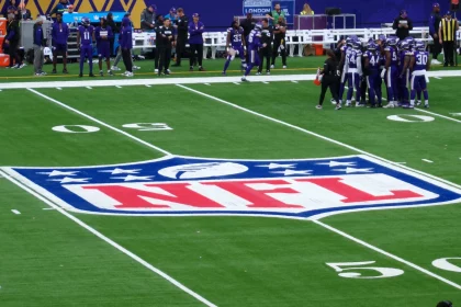FootballR - NFL Spiel - Footballspieler in lila Trikots versammeln sich auf dem Feld nahe der 50-Yard-Linie. In der Mitte des Spielfelds ist ein auffälliges NFL-Logo aufgemalt, fast wie ein künstlerischer Entwurf mit automatischer Präzision. Trainer und zusätzliche Spieler sind an der Seitenlinie zu sehen, im Hintergrund sind Stadionsitze und eine Anzeigetafel zu sehen. Diese Beschreibung wurde automatisch generiert.
