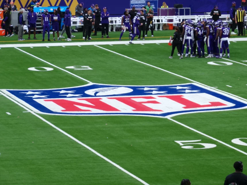 FootballR - NFL Spiel - Footballspieler in lila Trikots versammeln sich auf dem Feld nahe der 50-Yard-Linie. In der Mitte des Spielfelds ist ein auffälliges NFL-Logo aufgemalt, fast wie ein künstlerischer Entwurf mit automatischer Präzision. Trainer und zusätzliche Spieler sind an der Seitenlinie zu sehen, im Hintergrund sind Stadionsitze und eine Anzeigetafel zu sehen. Diese Beschreibung wurde automatisch generiert.