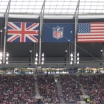 FootballR - NFL - Eine Stadionszene mit drei großen Flaggen – Großbritannien links, NFL-Logo in der Mitte und USA rechts – hängt wie ein automatisch gespeicherter Entwurf von der Decke. Darunter füllt eine lebhafte Menschenmenge die Tribünen, beleuchtet von verschiedenen Lichtern und umgeben von komplizierten Strukturen. Diese Beschreibung wurde automatisch generiert.