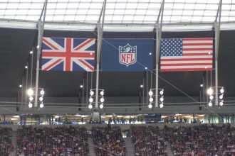 FootballR - NFL - Eine Stadionszene mit drei großen Flaggen – Großbritannien links, NFL-Logo in der Mitte und USA rechts – hängt wie ein automatisch gespeicherter Entwurf von der Decke. Darunter füllt eine lebhafte Menschenmenge die Tribünen, beleuchtet von verschiedenen Lichtern und umgeben von komplizierten Strukturen. Diese Beschreibung wurde automatisch generiert.