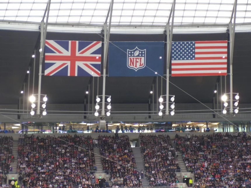 FootballR - NFL - Eine Stadionszene mit drei großen Flaggen – Großbritannien links, NFL-Logo in der Mitte und USA rechts – hängt wie ein automatisch gespeicherter Entwurf von der Decke. Darunter füllt eine lebhafte Menschenmenge die Tribünen, beleuchtet von verschiedenen Lichtern und umgeben von komplizierten Strukturen. Diese Beschreibung wurde automatisch generiert.