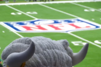 FootballR - NFL - Ein flauschiges Maskottchen mit großen gebogenen Hörnern überblickt ein Footballfeld mit dem NFL-Logo in der Mitte, ganz wie ein automatisch gespeicherter Entwurf der Aufregung. Das Gras ist leuchtend grün und Yard-Linien sind sichtbar. Die Zuschauer im Hintergrund sind verschwommen und konzentrieren sich auf das Maskottchen und das Logo. Diese Beschreibung wurde automatisch generiert.