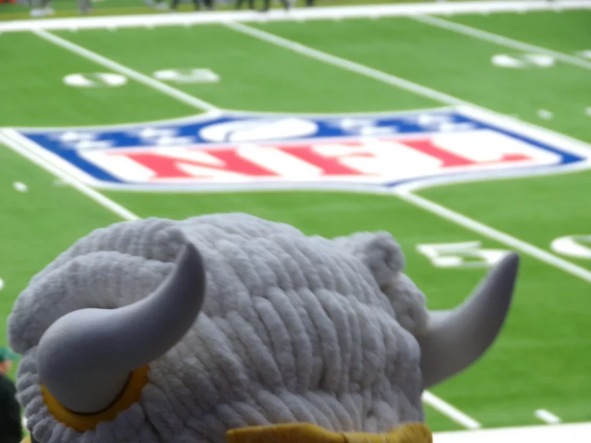 FootballR - NFL - Ein flauschiges Maskottchen mit großen gebogenen Hörnern überblickt ein Footballfeld mit dem NFL-Logo in der Mitte, ganz wie ein automatisch gespeicherter Entwurf der Aufregung. Das Gras ist leuchtend grün und Yard-Linien sind sichtbar. Die Zuschauer im Hintergrund sind verschwommen und konzentrieren sich auf das Maskottchen und das Logo. Diese Beschreibung wurde automatisch generiert.